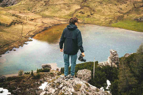 Person standing on a rock overlooking a serene lake and greenery, holding a camera.