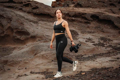 Woman in athletic wear walking on rocky terrain, holding a camera, showcasing an active lifestyle.