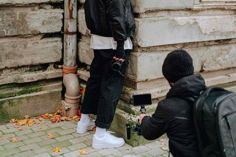 Photographer filming a model holding a camera against an urban backdrop with autumn leaves on the ground.