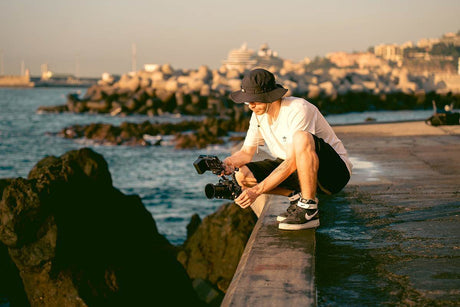 Photographer sitting on a pier with a camera, capturing images near rocky coastline at sunset.