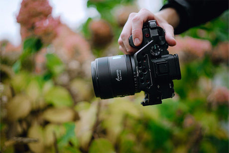 A person holding a camera with a lens in a garden, showcasing the camera's design and lens features.