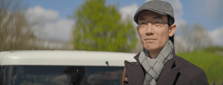 Person wearing a cap and scarf, standing next to a vehicle with a clear sky and greenery in the background.