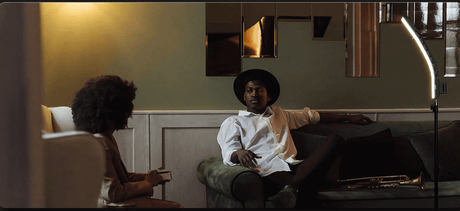 A jazz musician in a hat relaxes on a sofa while a woman listens, bathed in warm light.