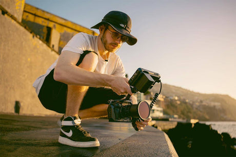 Person adjusting camera equipment while squatting by the ocean during sunset.