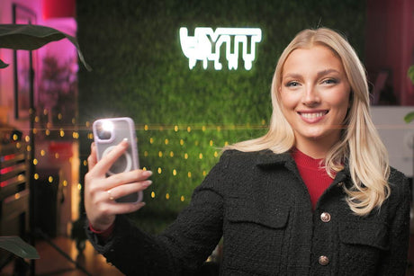 A smiling woman taking a selfie in a stylish setting with greenery and warm lighting.