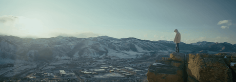 Person standing on a cliff overlooking a snowy mountain landscape at sunset.