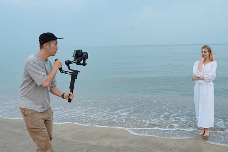 Photographer using a gimbal to capture video of a model in a white dress on a beach.