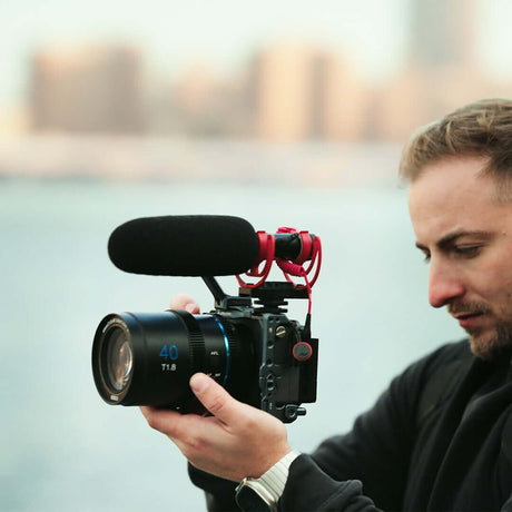 Man holding a camera with an autofocus lens and microphone, capturing video outdoors.