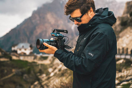 Man wearing sunglasses holding a camera outdoors in a mountainous landscape, exploring contemporary photography.