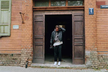 Person standing in a doorway, holding a camera, wearing a leather jacket in an urban setting.