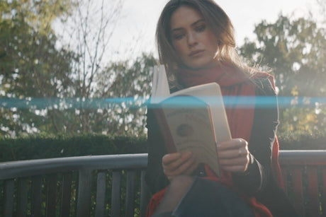 Woman reading a book outdoors on a bench, with sunlight filtering through trees, creating a serene atmosphere.