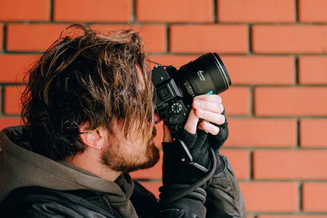 Man holding a camera, capturing a photo against a brick wall background, showcasing photography passion.