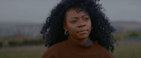 Woman with curly hair wearing a brown sweater, standing outdoors, with a thoughtful expression.