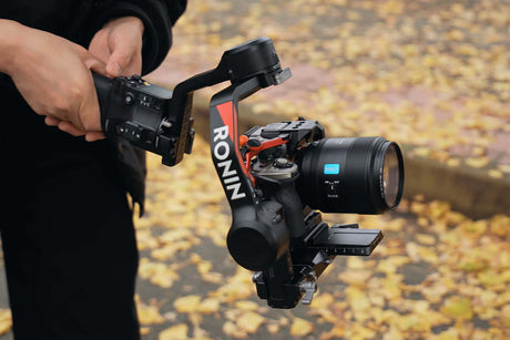 Person holding a Ronin gimbal stabilizer with a camera and lens, surrounded by autumn leaves.