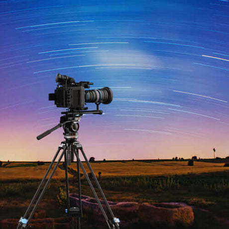 Camera mounted on a tripod capturing star trails in a twilight sky over a field.