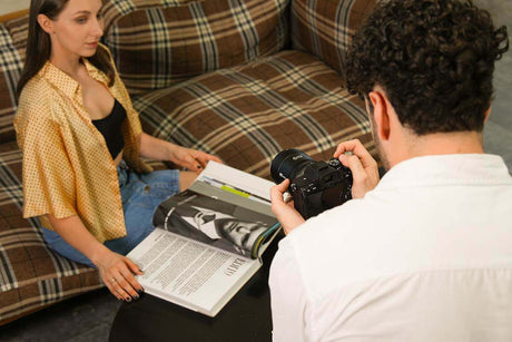 A photographer capturing a model sitting on a couch, reviewing a magazine, in a cozy home setting.