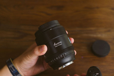 Hand holding an Aurora full frame camera lens on a wooden surface with accessories in the background.